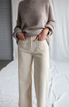 a woman standing on top of a bed wearing white pants and a brown turtle neck sweater