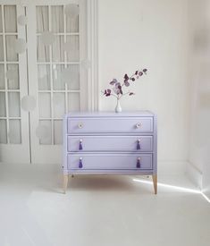 a white room with a purple dresser and flowers in a vase on the top shelf