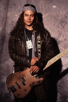 a man with long hair holding a bass guitar in front of a gray wall and wearing a bandana