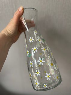 a hand holding a glass vase with white and yellow daisies on the bottom, in front of a wall