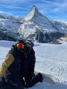 two snowboarders sitting in the snow with their skis on and mountains in the background