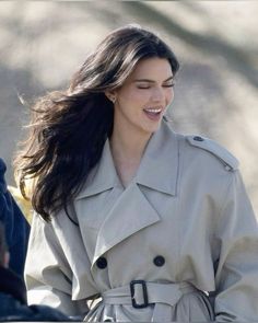 a woman in a trench coat smiles as she walks down the street with her hand on her hip