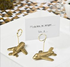 two gold airplane key chains are on a white plate with a place card in the middle