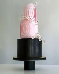 a three tiered pink and black cake with white flowers on the top, sitting on a table