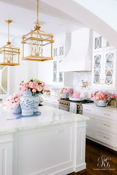 a white kitchen with pink flowers in a vase on the counter and hanging lights above it