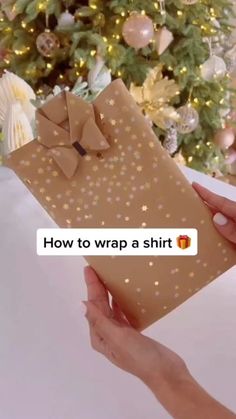 someone holding a wrapped gift in front of a christmas tree that says how to wrap a shirt