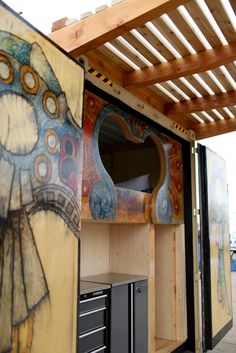 an outdoor kitchen area with cabinets and artwork on the wall, under a pergolated roof