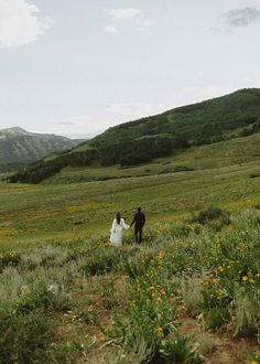 a bride and groom are walking through a field with wildflowers in the foreground