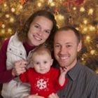 a man and woman holding a baby in front of a christmas tree with lights on it