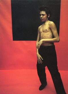 a man with no shirt standing in front of a red background wearing black pants and holding his hand on his hip