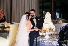 a bride and groom standing in front of their wedding cake at the reception table with guests