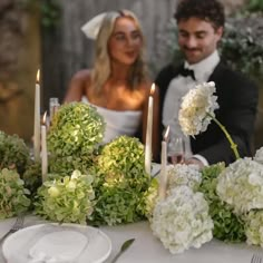 a man and woman sitting at a table with flowers in front of them, surrounded by candles