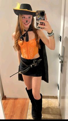 a woman in a cowboy hat is taking a selfie with her cell phone while wearing an orange shirt and black skirt