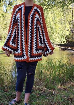 a woman standing next to a river wearing a crocheted jacket