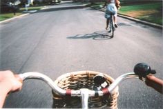 two people riding bikes down a street with houses in the background and grass on both sides