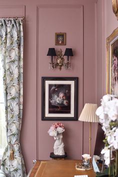 a room with pink walls and white flowers on the desk in front of a window