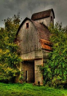 an old barn in the middle of a field with trees around it and dark clouds overhead