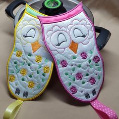 two oven mitts are decorated with flowers and an owl face on them, one has a pink ribbon around its neck