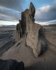 a large rock formation in the middle of a barren area with mountains and clouds behind it