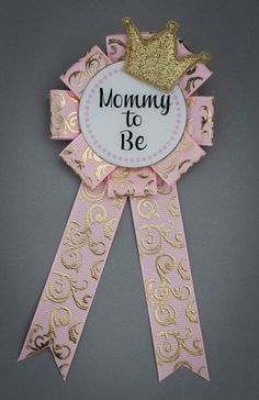 a pink ribbon with a gold crown on top and the words mommy to be above it