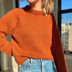 a woman wearing an orange sweater and denim skirt standing in front of a window with her hands on her hips