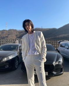 a man standing in front of some cars wearing a white suit and black shoes with his hands on his hips