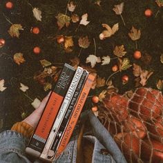 a person is holding three books in their hands while laying on the ground with autumn leaves