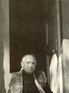 an old man wearing a fur vest sitting on a window sill with his hands in his pockets