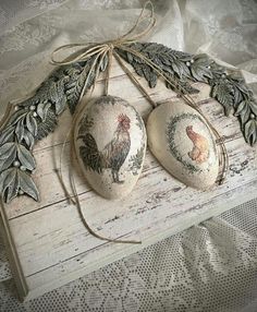 two decorative rocks with roosters on them are tied to a white wooden board and decorated with leaves