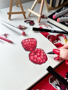 a person drawing raspberries on paper with markers and pencils next to them