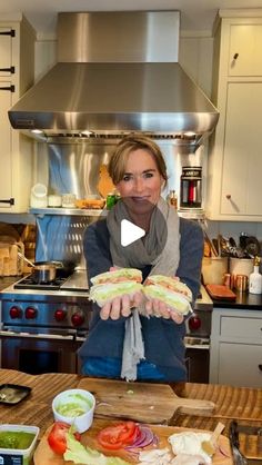 a woman holding sandwiches in her hands while sitting at a kitchen table with food on it