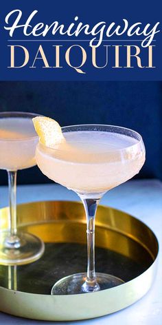 two margaritas sitting on top of a metal tray with lemon wedged in the rim