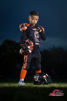 a young boy wearing an orange and black uniform holding a football helmet while standing in the grass