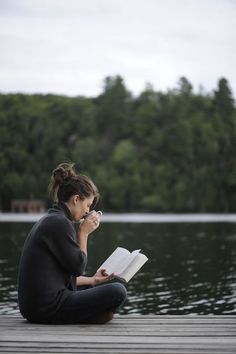 a woman sitting on a dock reading a book and looking at the camera while holding her hand to her face