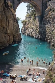 people are on the beach in front of an old stone bridge and blue water under it