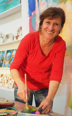 a woman in red shirt painting on canvass with brush and paintbrush at table