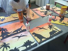 a man is painting dinosaurs on paper at a table with paint cans and other items