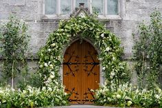 a wooden door surrounded by white flowers and greenery in front of a stone building