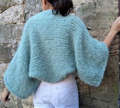 a woman standing in front of a stone wall wearing a blue knitted sweater and white shorts