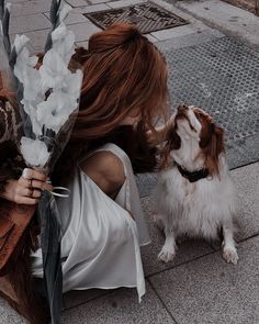 a woman holding a bouquet of white flowers next to a brown and white dog on the sidewalk