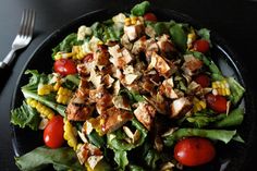 a salad with chicken, corn and tomatoes on a black plate next to a fork