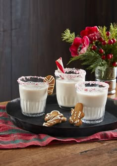 three glasses filled with milk and cookies on a black platter next to red flowers