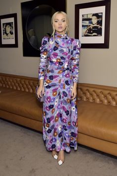 a woman in a purple dress standing next to a brown leather couch with pictures on the wall behind her