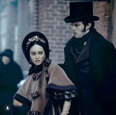 a man and woman dressed in period costumes walking down the street together, with one wearing a top hat