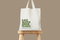 a white tote bag sitting on top of a wooden stool with the words save water drink margs printed on it