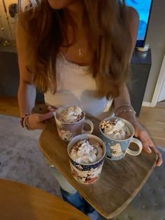 a woman holding two cups of coffee on top of a wooden tray with whipped cream in them