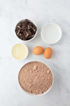 ingredients for chocolate cake laid out in bowls on a marble countertop, including eggs and flour