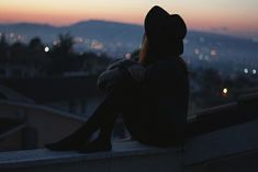 a woman sitting on top of a roof looking at the city lights in the distance