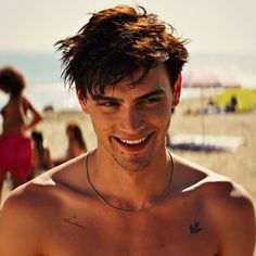 a man with no shirt on standing in front of the beach and smiling at the camera