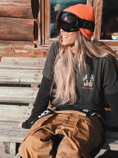 a woman sitting on a wooden bench wearing goggles and an orange beanie hat